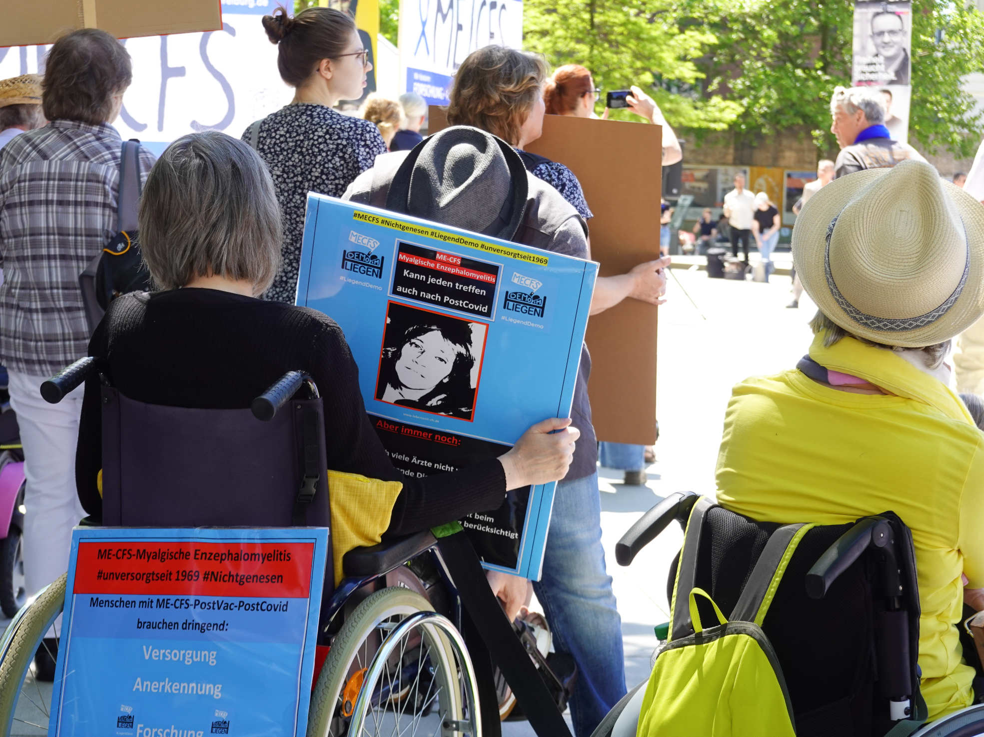 Bildbeschreibung: Foto mit zwei Demonstrantinnen im Rollstuhl. Eine der beiden hat zwei Schilder, die über die katastrophale Lage von ME/CFS aufklären und Versorgung, Anerkennung und Forschung fordern.